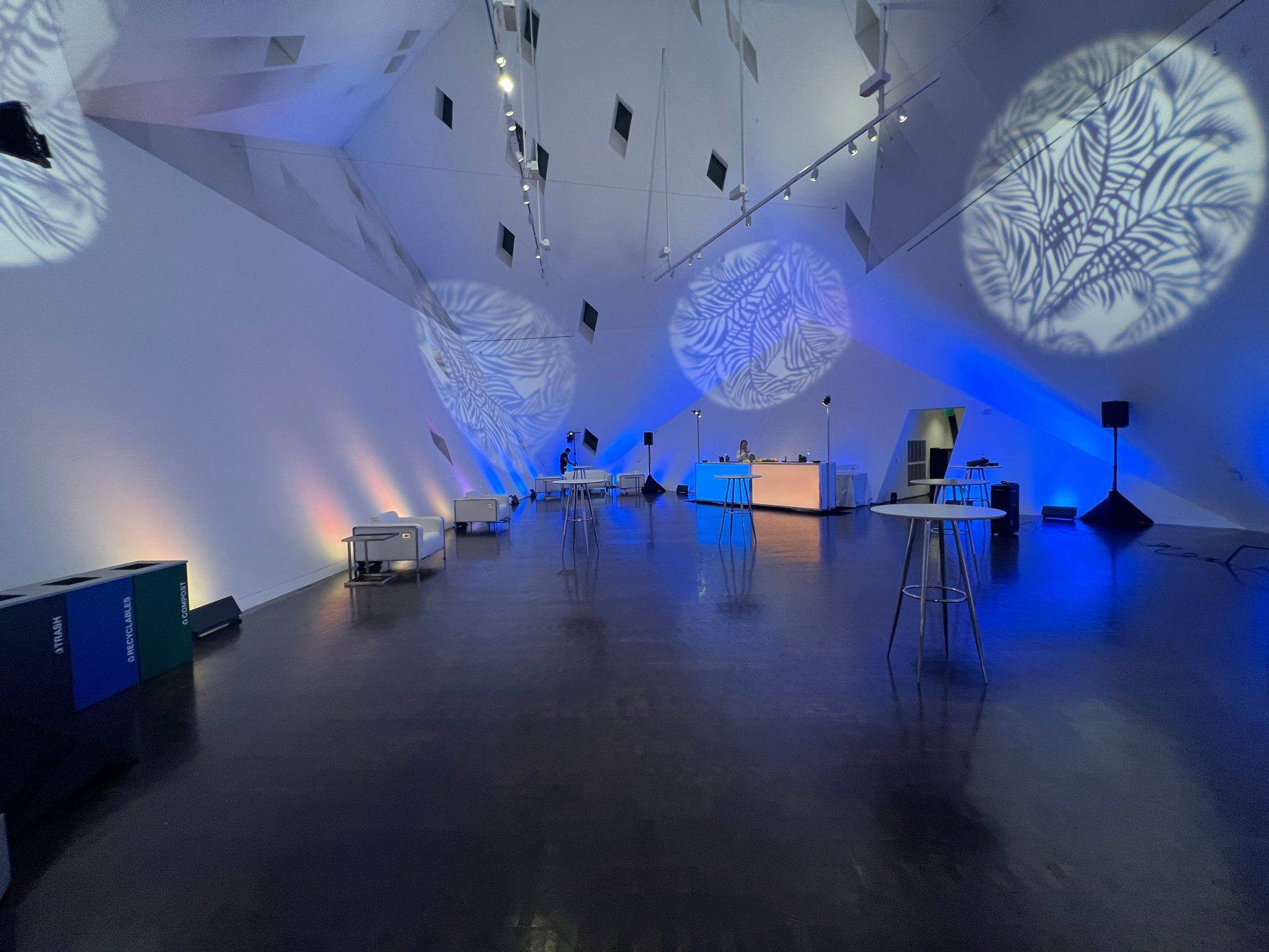 White gallery room with palm-leaf gobo patterns on the walls and blue uplighting